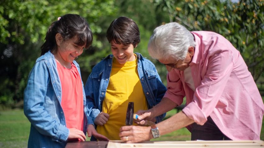 Grandfather teaching grandchildren woodworking skills outdoors on a sunny day.